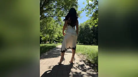 CassyTorres stands on a stone path in a park, wearing a white lace-trimmed slip dress and white high heels, looking back over her shoulder.