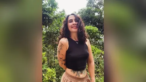 RossiNoleen smiles wearing a black tank top and tan bottoms, showing off her tattoos and curly hair against a lush green forest background.