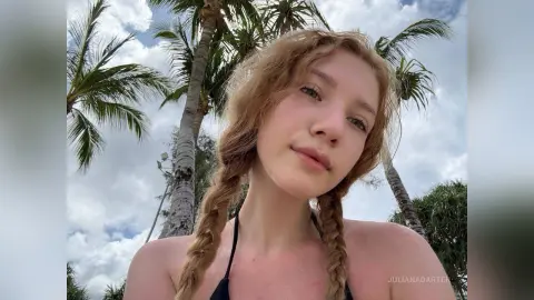 Smiling young woman with braided hair in a black bikini under palm trees on a sunny tropical day.