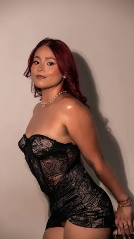 Confident woman in a black lace strapless dress posing against a neutral wall in soft studio lighting.
