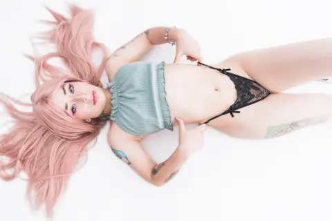Young woman with long pink hair in a sage green crop top and black lace bikini lying on a white background.