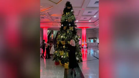 Elegant woman in a black dress standing by a large glowing Christmas tree in a festive luxury hotel lobby.