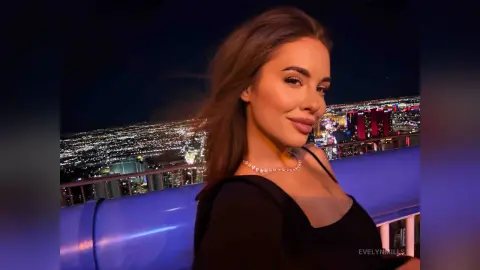 EvelynMills smiles while wearing a black top against a bright night view of the Las Vegas strip.