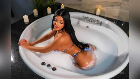 Relaxed woman in a blue bikini relaxing in a white bathtub surrounded by glowing candles in a luxury bathroom.