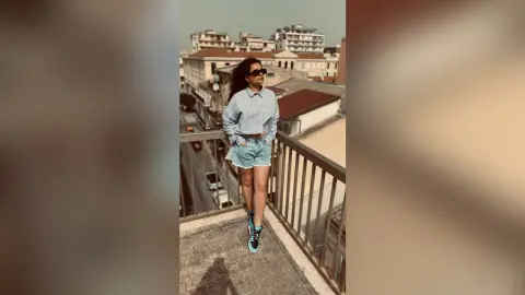 Stylish woman in a light blue shirt and denim shorts standing on a city balcony under bright sunlight.