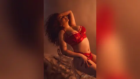 Woman in red lace lingerie posing in warm, moody lighting for a sensual indoor portrait.