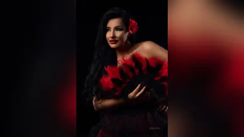 MissyJolie poses with long black hair and a red rose behind her ear, wearing a red and black feathered outfit against a black background.