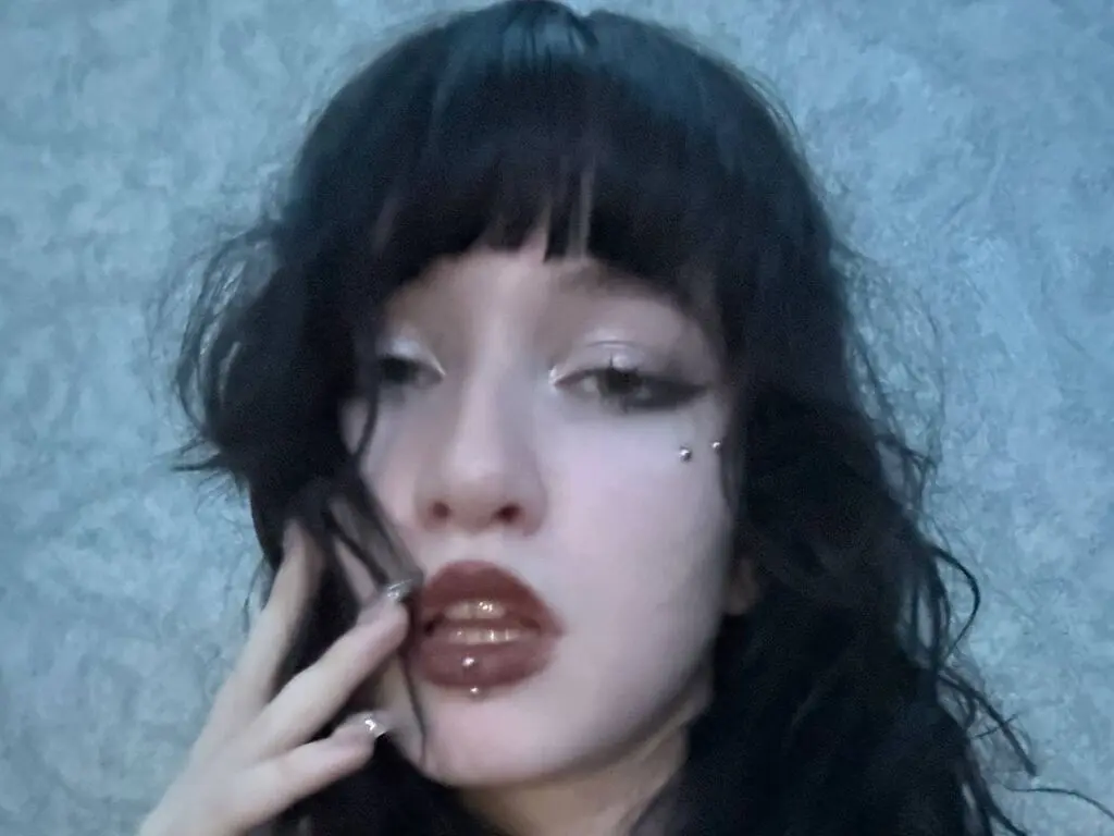 Young woman with dark bangs and winged eyeliner posing with a moody, aesthetic expression against a textured wall.