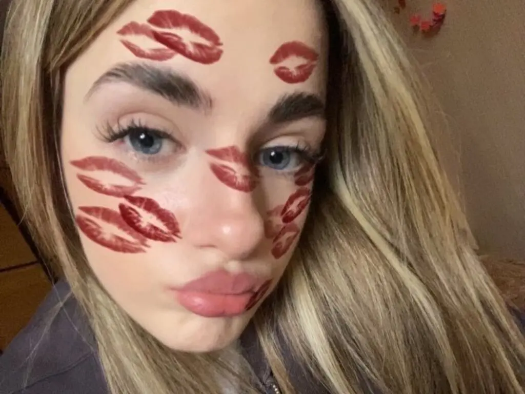 Close-up portrait of a blonde woman with red lipstick kiss marks on her face in a soft, playful mood.