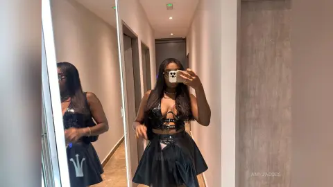 Young woman in a black leather corset and mini skirt taking a mirror selfie in a modern hallway.