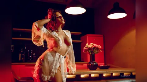 Rebecca poses in a white lace robe in a dimly lit kitchen, showing off her breasts and hair.