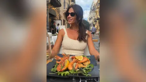 AnaMathers smiles wearing sunglasses and a white tank top while sitting at an outdoor table with a large seafood platter.