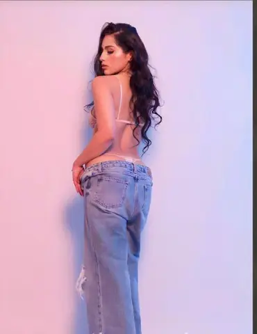 Young woman in denim jeans and white lingerie posing against a pastel pink and blue studio background.
