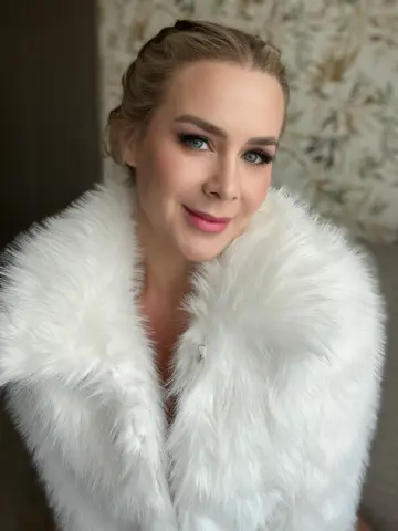 Elegant woman in a white faux fur coat with glamorous makeup posing in a bright, soft-lit setting.