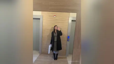 Stylish woman in a black coat and grey dress taking a mirror selfie in a modern wood-paneled elevator.