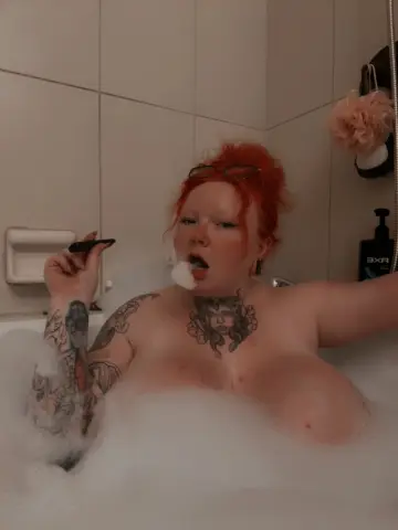 RaelynnRiley poses in a white bubble bath, showing her red hair and chest tattoos while holding a piece of white foam in her mouth.
