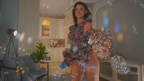 Smiling woman in a floral print dress dancing amidst floating bubbles in a bright, modern living room.