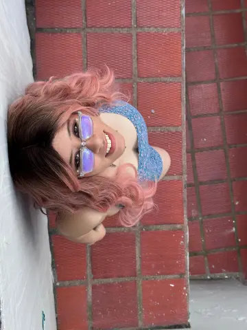 Izzumi smiles wearing light blue lace lingerie and silver-rimmed glasses while leaning against a white wall over red floor tiles.