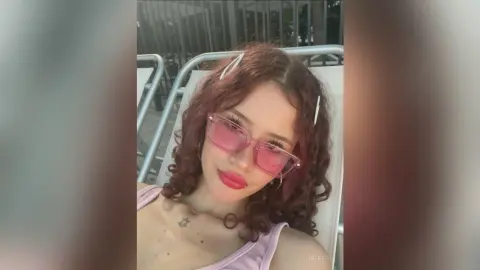 Young woman with curly red hair and pink sunglasses posing outdoors in a casual summer outfit.