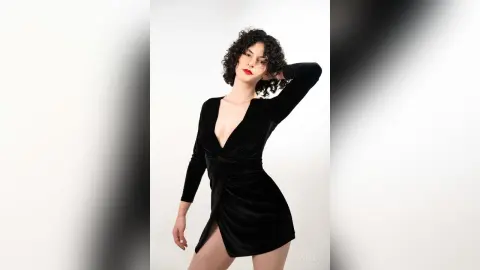 RoxyGraff poses in a low-cut black velvet dress with long sleeves against a white background, showing her dark curly hair and red lipstick.