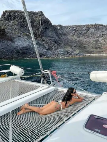 Woman in a bikini relaxing on a catamaran net near rocky coastal cliffs under a clear sky.