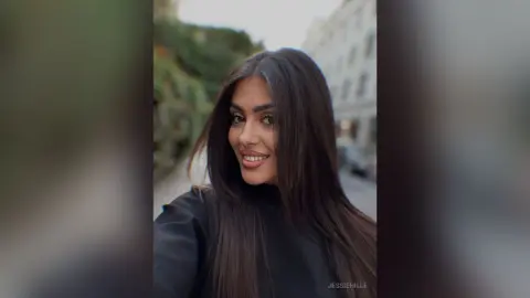Smiling brunette woman in a black turtleneck outfit posing outdoors in a city setting.