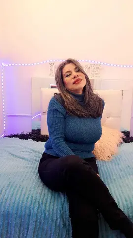 Smiling woman in a blue turtleneck sweater sitting on a bed in a cozy bedroom with fairy lights.