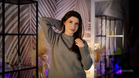 Smiling young woman in a cozy grey sweater posing in a modern room with tropical leaf wallpaper.