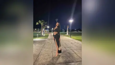 NicoleBella stands outside at night wearing a black crop top and a long black skirt with a high slit, showing her bare leg.