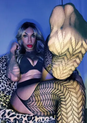 Tattooed woman in black lingerie and patterned fishnet tights relaxing on a leopard print chair in moody blue light.