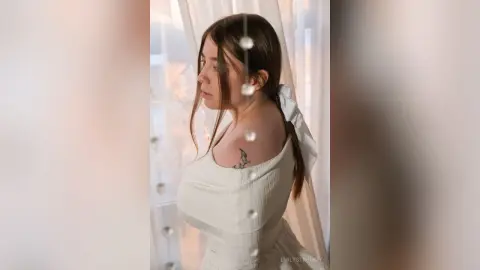 Young woman in a white summer dress with a hair bow looking out a window in soft, dreamy lighting.