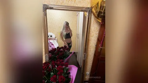 Blonde woman in a black crop top posing with red roses in a mirror selfie for a romantic celebration.