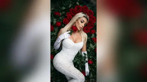 Elegant blonde woman in a white lace dress and gloves posing against a romantic red rose floral background.