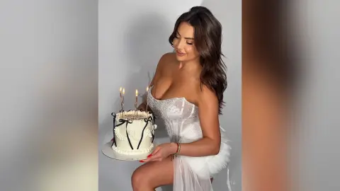 Smiling woman in a white strapless dress holding a lit birthday cake in a bright indoor setting.