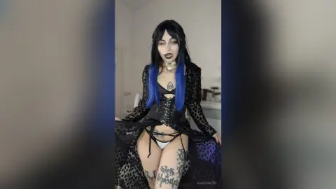 Gothic woman with blue hair in black lace corset and sheer robe posing in a moody bedroom setting.