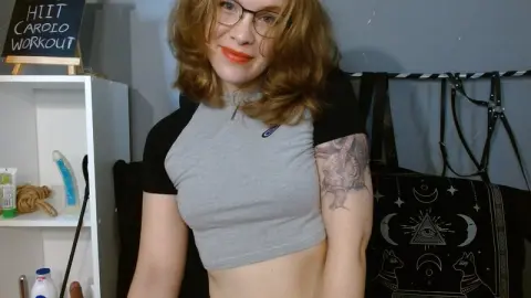ChillBillie wears glasses and a grey crop top while smiling at the camera in her room.