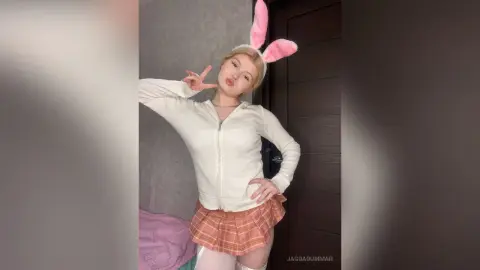 Cheerful young woman in a white zip-up hoodie and plaid mini skirt wearing pink bunny ears indoors.