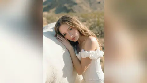 Dreamy portrait of a blonde woman in a white off-the-shoulder dress leaning against a white horse in a desert setting.