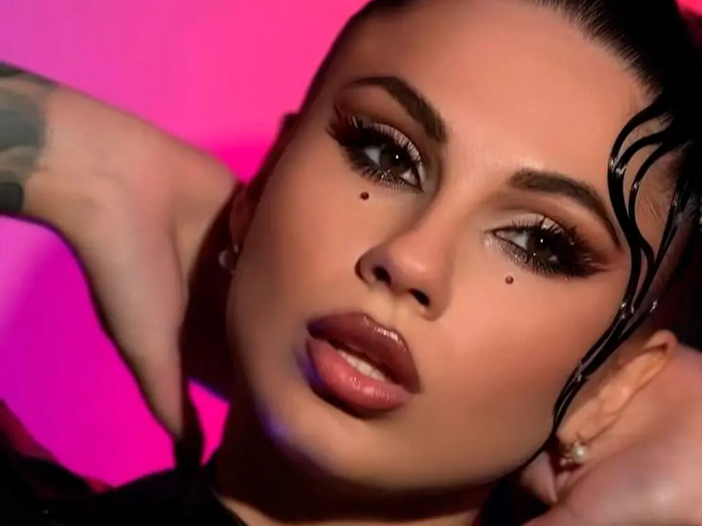 Glamorous woman with dramatic eye makeup and dark hair posing against a vibrant pink background.