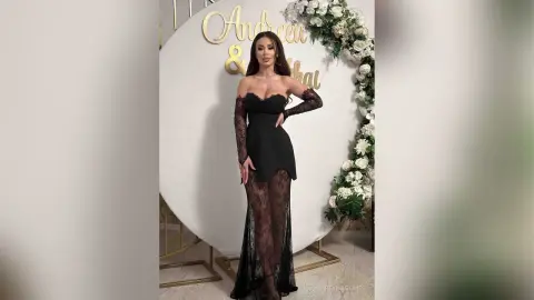 ReynaGomes poses in a long black off-the-shoulder lace dress with long sleeves, standing in front of a white circular backdrop decorated with white flowers.