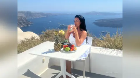 MissyJolie sits at a white table overlooking the sea, wearing a white top and holding a white mug.