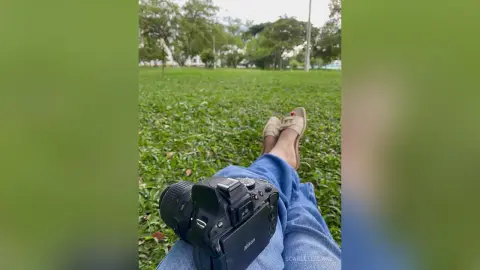 First-person view of a person in blue jeans relaxing on green grass with a Nikon DSLR camera.