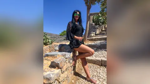 MissyJolie stands outdoors in a black lace dress and sunglasses, posing against a stone wall under a clear blue sky.