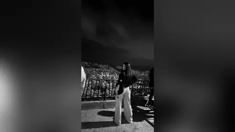 Stylish woman in wide-leg trousers and black top standing on a balcony overlooking a scenic mountain landscape.