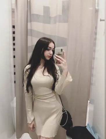 Young woman with long dark hair wearing a beige long-sleeve mini dress taking a mirror selfie in a bright room.