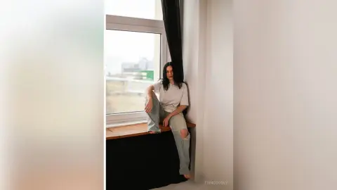Young woman in a white t-shirt and ripped jeans sitting on a window sill in a bright, minimalist room.