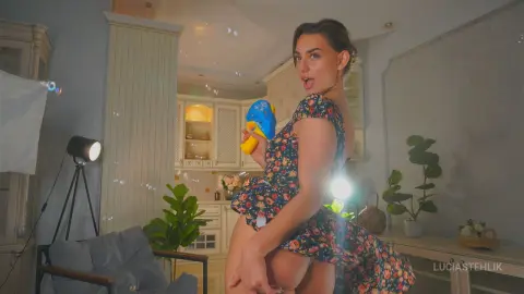 Cheerful woman in a floral summer dress holding a blue water gun in a bright, modern living room.