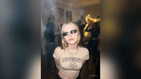 JaimieBez wears sunglasses and a tan t-shirt with a university logo while standing in a room with posters on the wall.