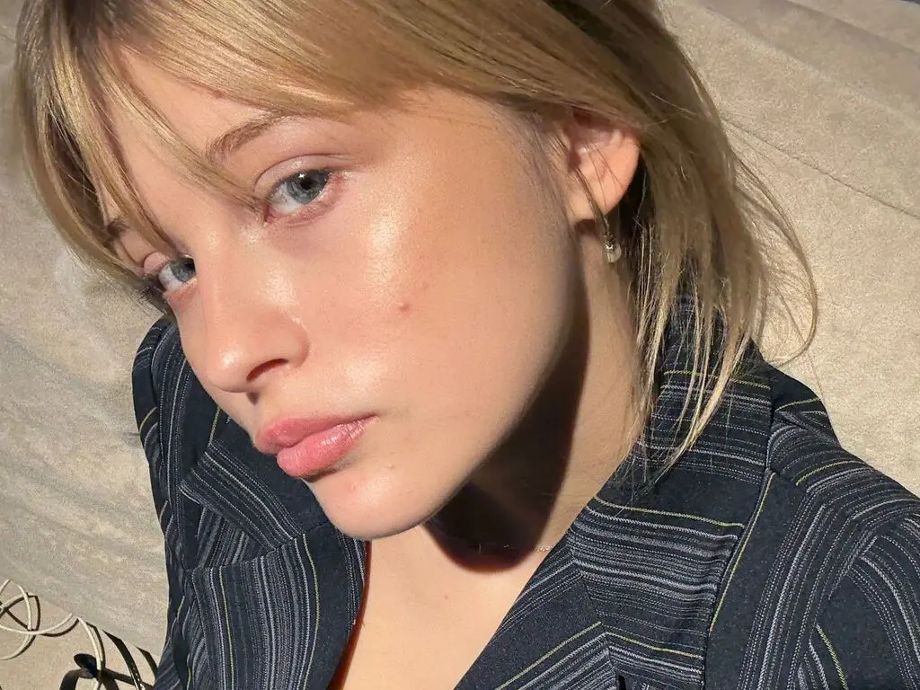 Close-up portrait of a blonde woman in a striped blazer enjoying warm sunlight in a serene outdoor setting.