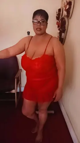Curvy woman in a red lace chemise posing in a cozy bedroom setting.